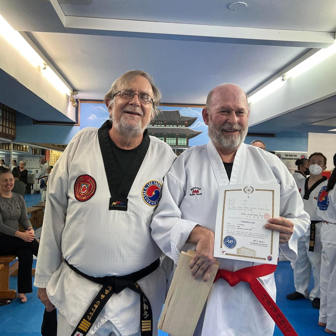 John-David Skene, wearing a red belt and holding a promotion certificate and a broken board, stands beside a black-belt instructor in a Taekwondo school.
